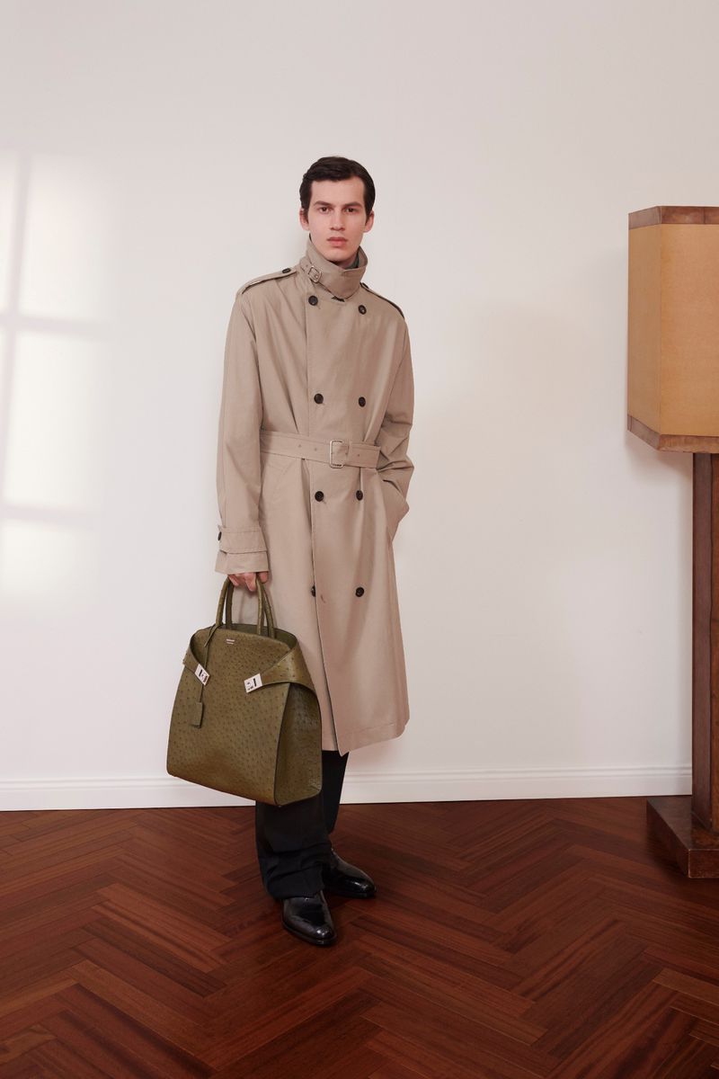 A stylish man in a beige trench coat holds a textured green leather bag, standing on a herringbone wood floor against a white wall.