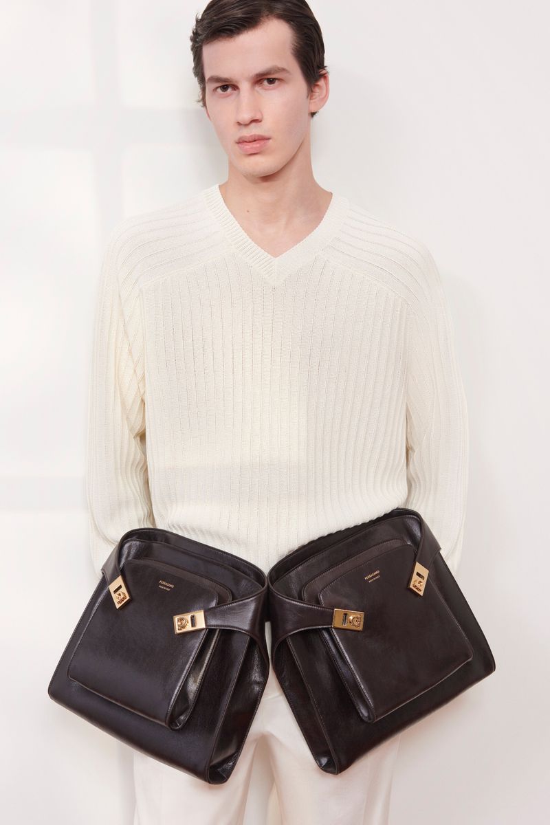 Fashion shot of a man wearing a white sweater with two black handbags hanging from his shoulders against a plain white background.