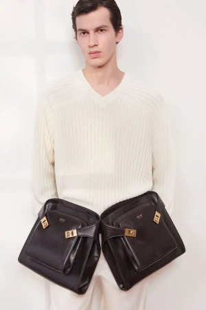 Fashion shot of a man wearing a white sweater with two black handbags hanging from his shoulders against a plain white background.