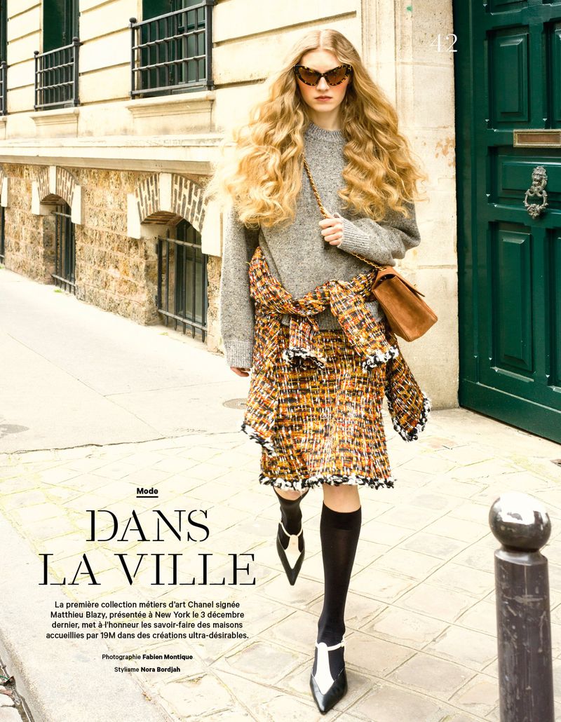 Model with voluminous blonde hair walking down a street wearing a grey sweater, patterned tweed skirt, and pointed heels.