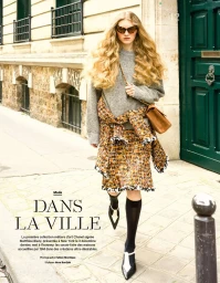 Model with voluminous blonde hair walking down a street wearing a grey sweater, patterned tweed skirt, and pointed heels.