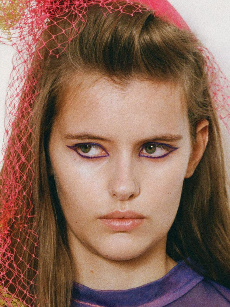 Close-up portrait of model Ruby Pritchett with graphic purple eyeliner and a pink mesh headpiece.