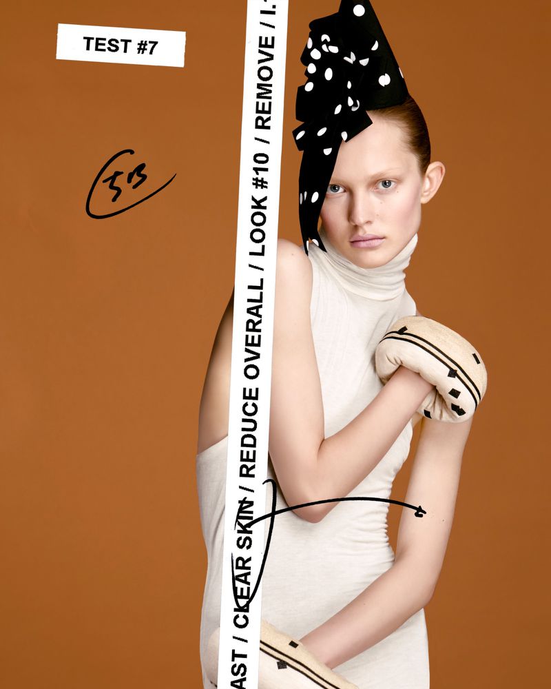 Model Viktoria Wirs wearing an off-white turtleneck dress and black polka-dot headpiece against a warm brown background.