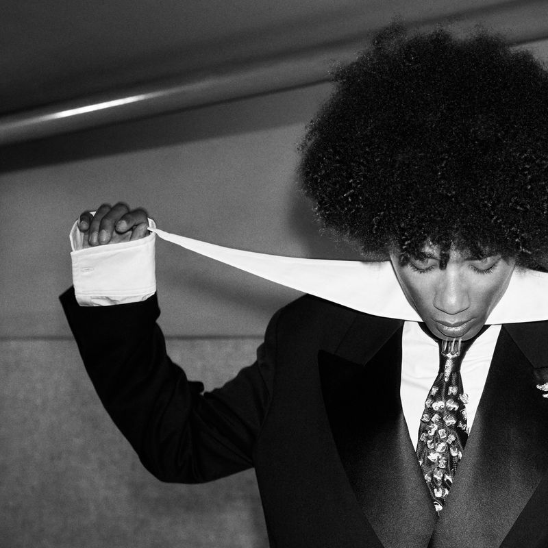 Black and white portrait of a man with an afro in a tuxedo holding a white collar ribbon across his neck.