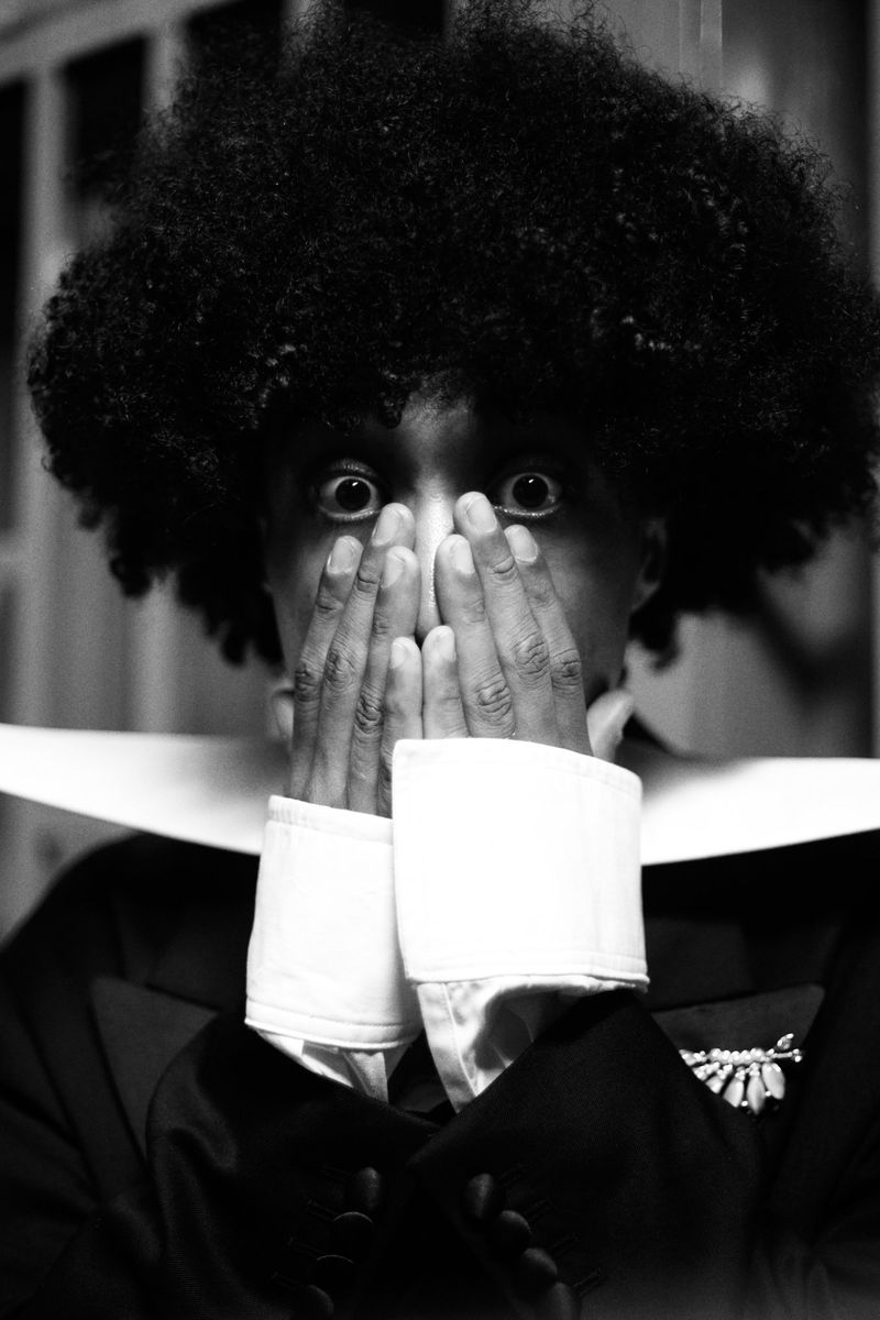 Black and white portrait of a person with an afro covering their mouth with hands, wearing wide white cuffs and collar.