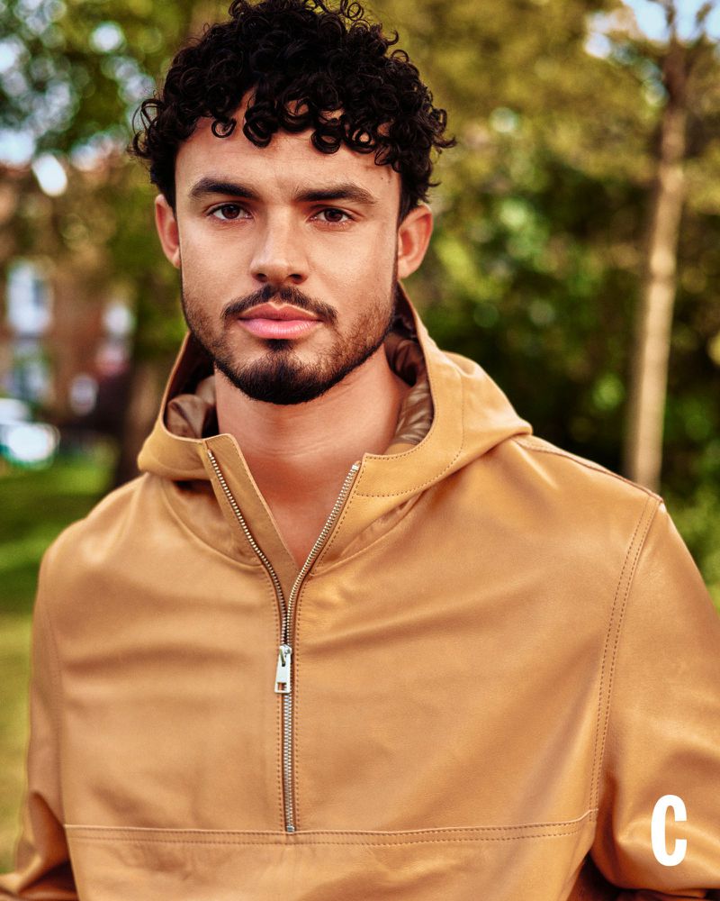 Matthew Broome posing outdoors in a camel hooded leather half-zip jacket against soft-focus trees.