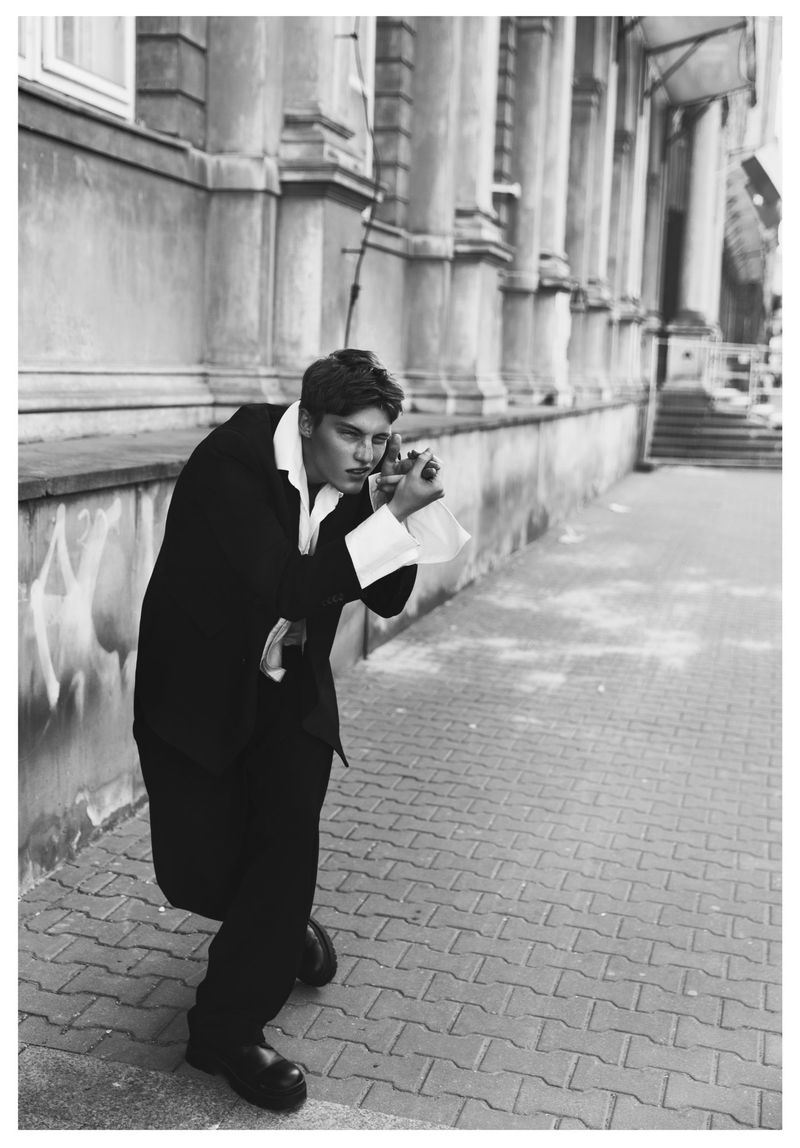 Black and white portrait of a man leaning against a classical stone building exterior wearing a dark suit