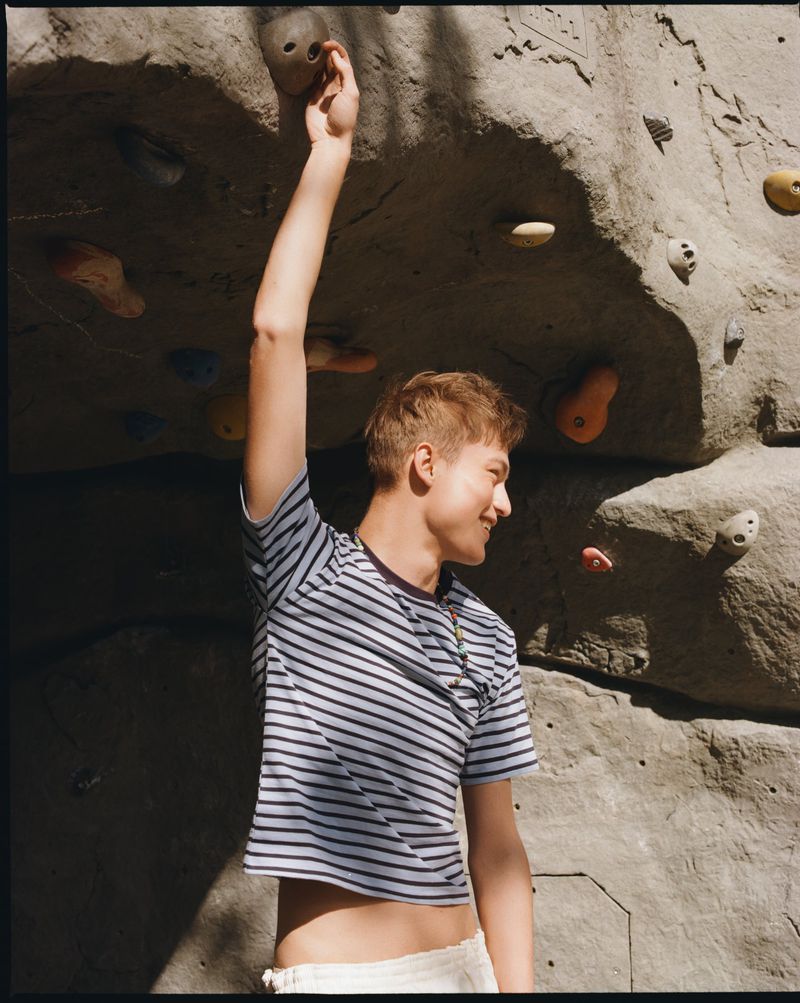 Dominik Datko reaching for a climbing hold on an artificial rock wall wearing a blue striped crop tee for Hube Magazine.