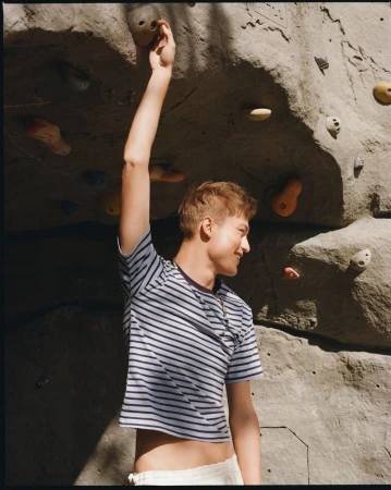 Dominik Datko reaching for a climbing hold on an artificial rock wall wearing a blue striped crop tee for Hube Magazine.