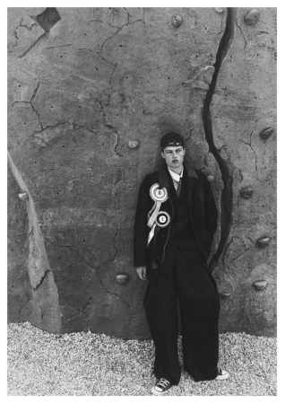Model Dominik Datko wearing an oversized black coat with prize ribbons and wide-leg trousers leaning against a climbing wall.