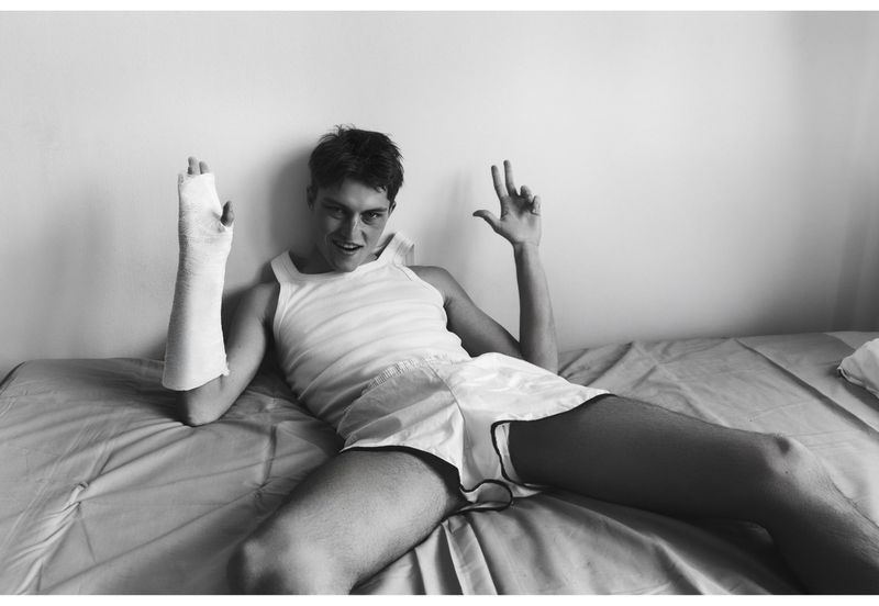 Model Dominik Datko wearing a white tank top, shorts, and an arm cast while reclining on a bed in black and white.