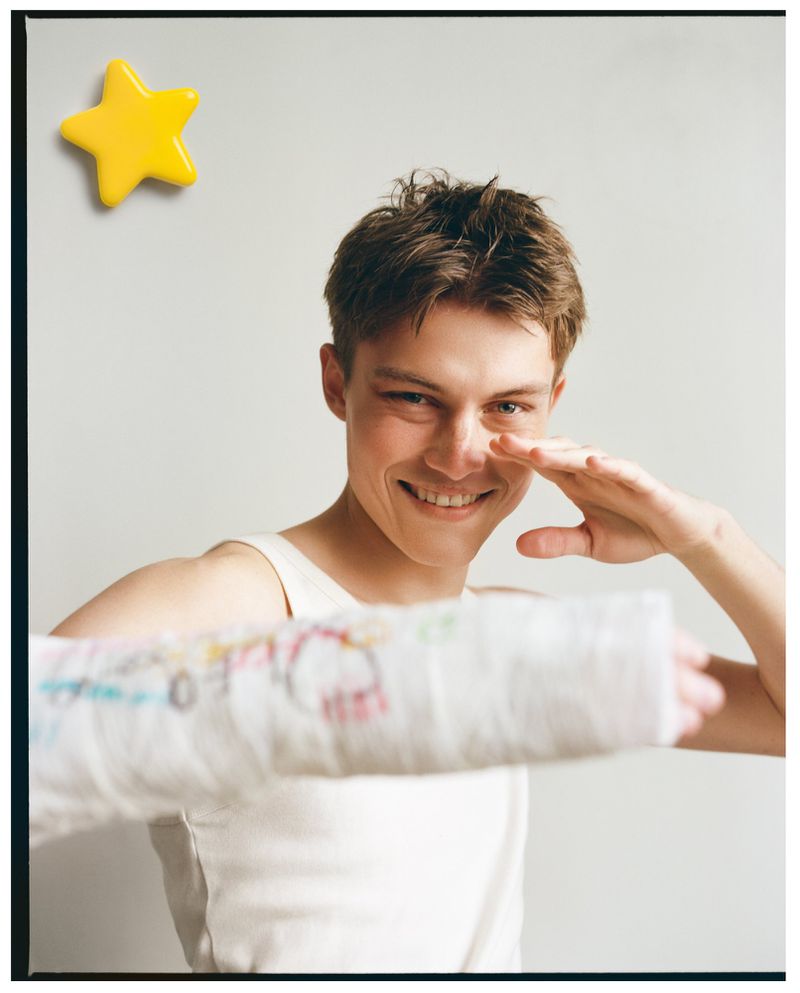 Dominik Datko smiling in a white tank top while holding out an arm in a decorated plaster cast against a neutral backdrop.