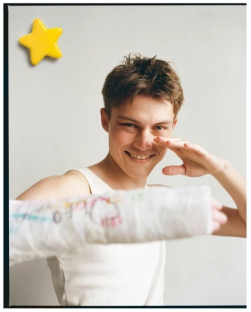 Dominik Datko smiling in a white tank top while holding out an arm in a decorated plaster cast against a neutral backdrop.