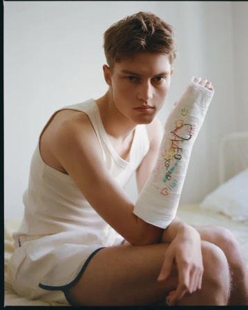 Young man in a white tank top sitting on a bed with a signed white cast on his arm for Hube Magazine LEO project.