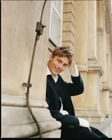Model Dominik Datko smiles while leaning outdoors against a stone building, wearing a black tailored suit and white collared shirt.