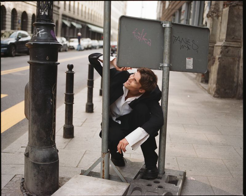 Model Dominik Datko crouching behind a metal street sign wearing an oversized black suit and white shirt on a city sidewalk.