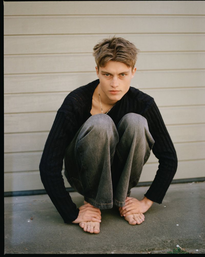Model Dominik Datko crouches barefoot in black knitwear and grey denim jeans against horizontal panel siding.
