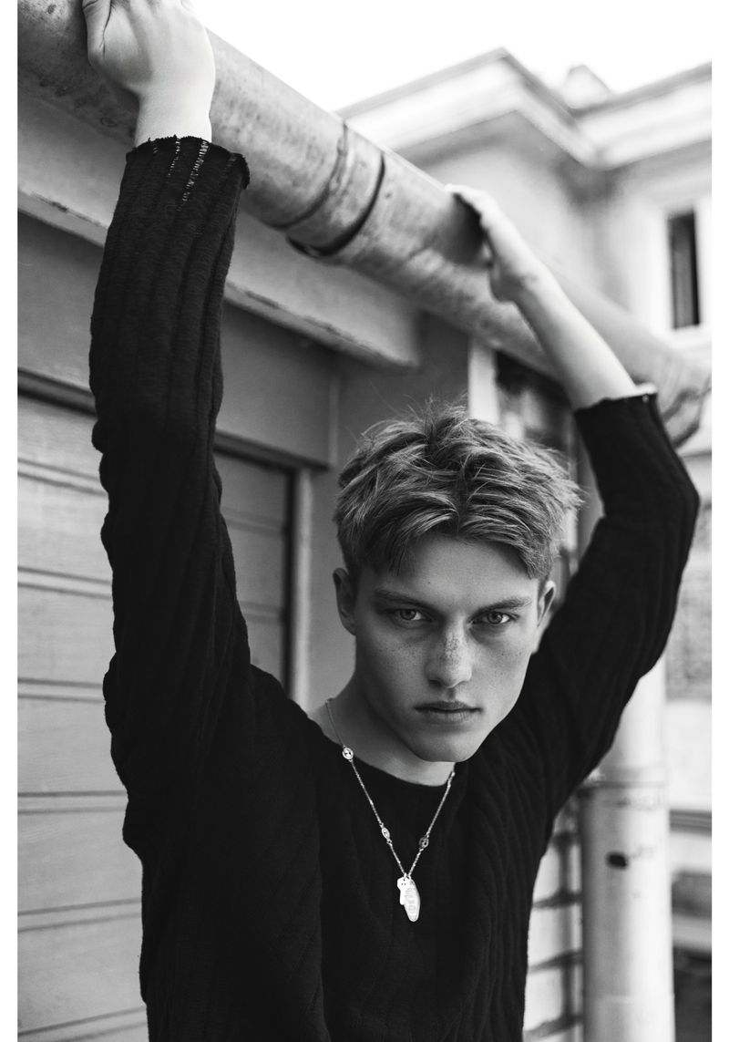 Dominik Datko posed in a dark textured sweater holding an overhead metal pipe against an urban exterior wall in black and white.