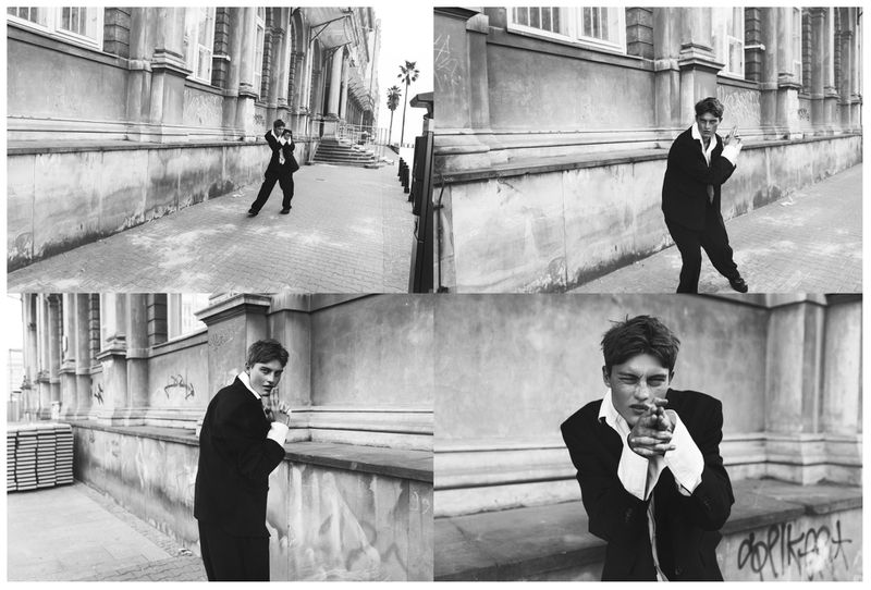 Four-panel black and white sequence of model Dominik Datko in an oversized black suit posing with finger guns by an exterior stone wall.