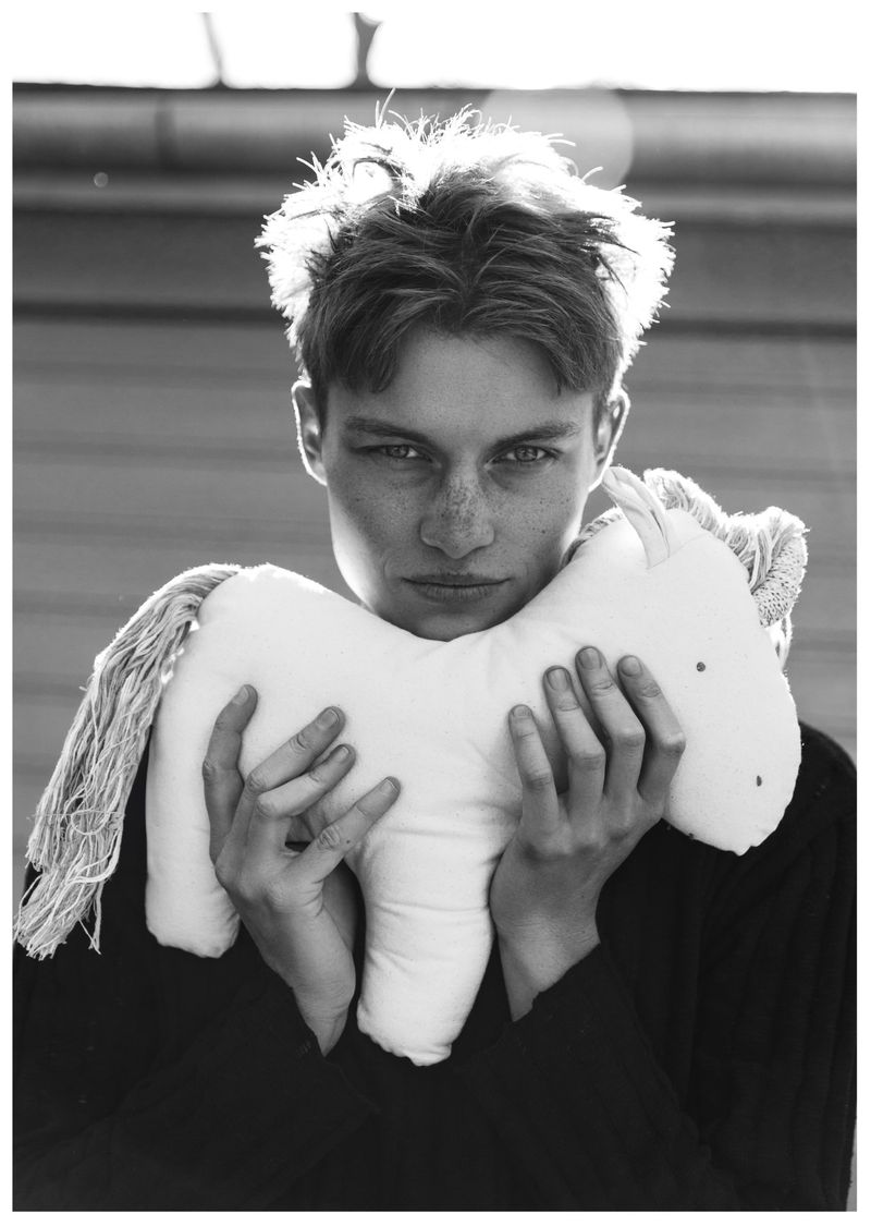 Close-up monochrome portrait of a young man holding a white stuffed toy horse against an outdoor wall.