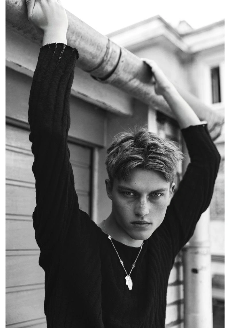 Dominik Datko posed in a dark textured sweater holding an overhead metal pipe against an urban exterior wall in black and white.