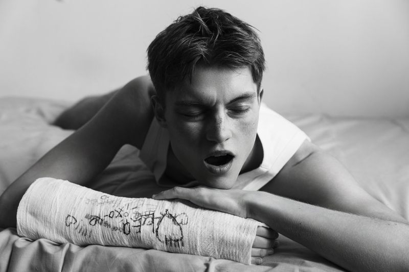 Model Dominik Datko yawning on a bed wearing a white tank top and a doodled arm cast in black and white.