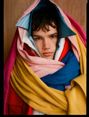Model Ian Jeffrey wrapped in multiple colorful layered shirts and knitted fabrics against a wooden backdrop.