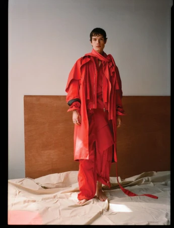 Full-length portrait of a male model wearing a vibrant red layered coat and matching red trousers standing in a studio.