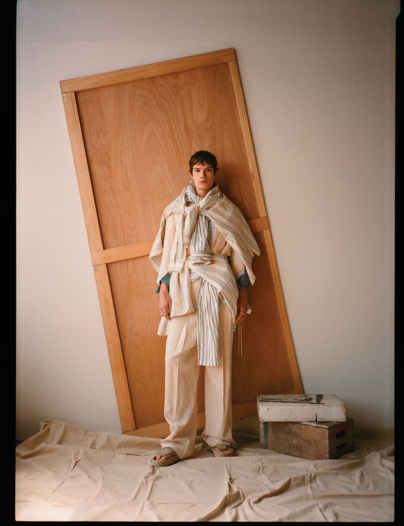 Model Ian Jeffrey wearing layered cream garments and wide-leg trousers standing before a leaning wooden board in a studio.