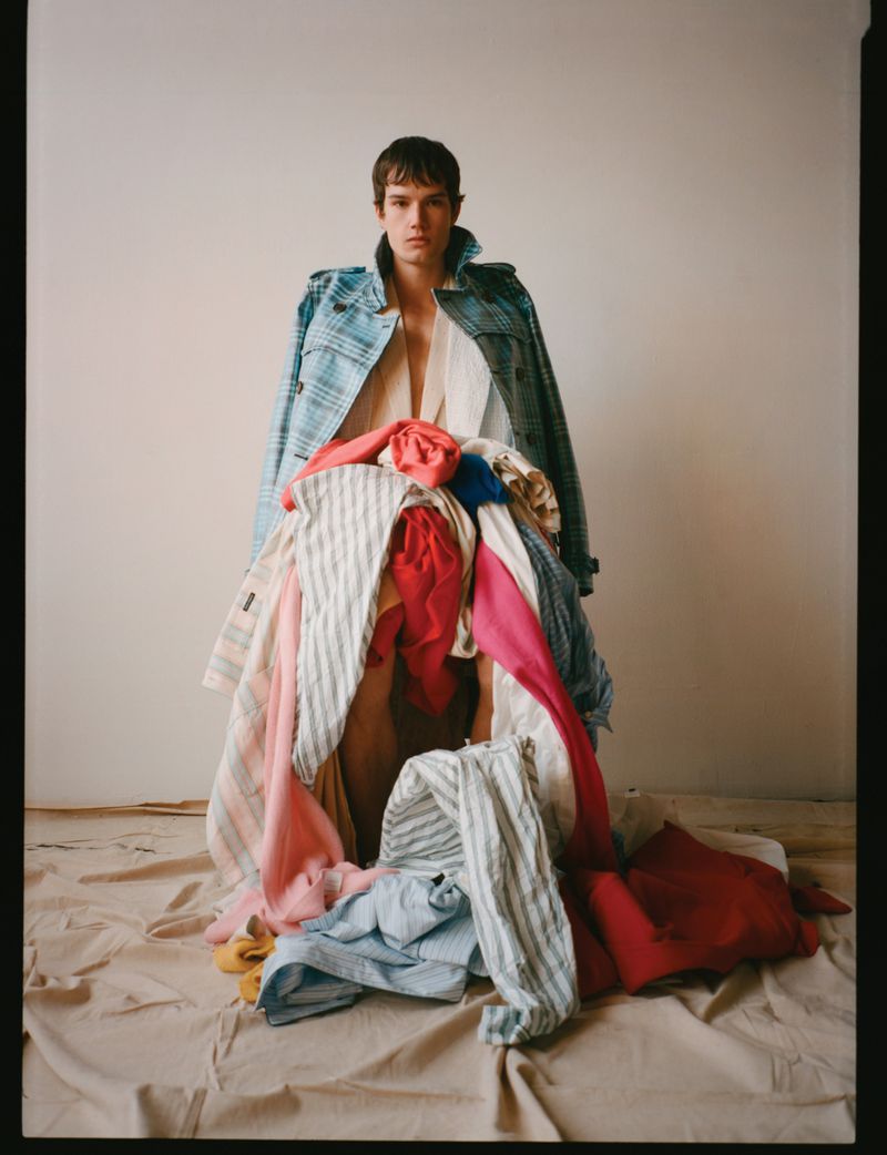Model Ian Jeffrey standing with a large pile of layered colorful clothing draped in front of him against a plain studio backdrop.