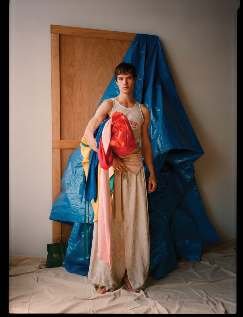 Model Ian Jeffrey wearing a white tank top and draped beige trousers holding colorful fabrics against a blue tarp backdrop.
