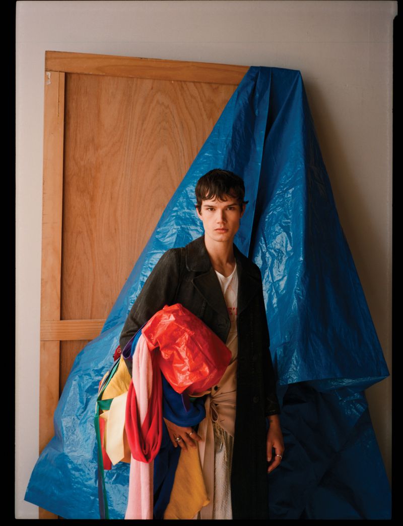 Model wearing a dark coat holding a bundle of colorful fabrics against a wooden panel and blue plastic sheet