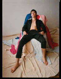 Model Ian Jeffrey sitting shirtless in an open black jacket and black pants on a chair draped with colorful fabrics.