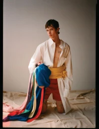 Model Ian Jeffrey kneeling on a canvas drop cloth wearing an open white shirt, yellow sash, and colorful cascading fabric ribbons.