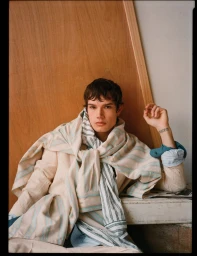 Model Ian Jeffrey seated in layered striped and beige garments leaning against a wooden board for an editorial shoot.