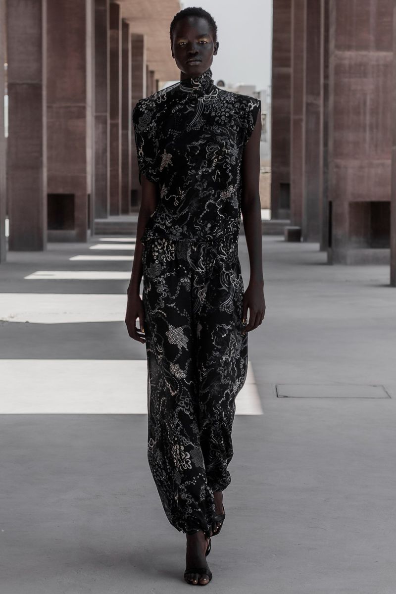 An adult female fashion model poses outdoors wearing a black and silver patterned jumpsuit. Pillars are in the background.