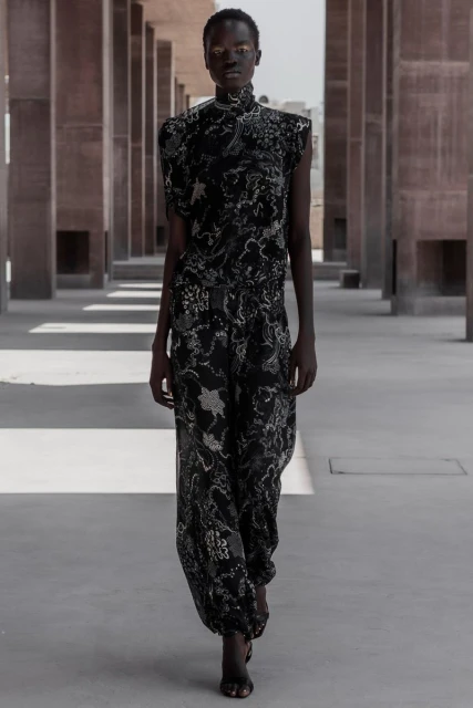 An adult female fashion model poses outdoors wearing a black and silver patterned jumpsuit. Pillars are in the background.