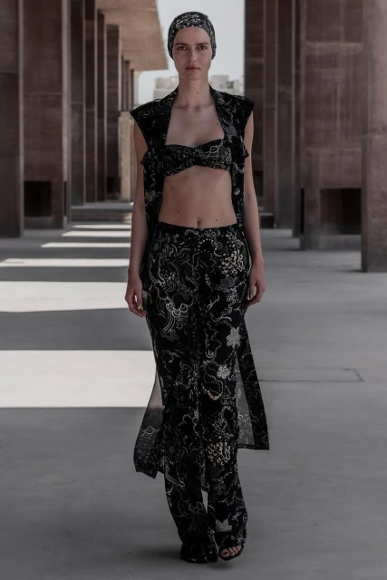 A woman walks in a flowing floral pantsuit in a colonnade, bathed in sunlight, creating a fashion-forward and visually striking image.