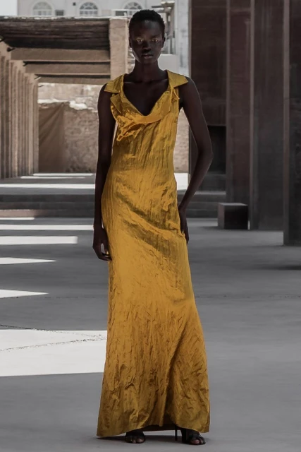 Fashion shot of a black female model in a long, golden gown posing in an outdoor setting with concrete structures and neutral tones.