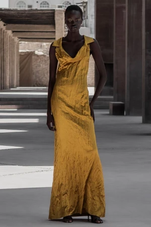 Fashion shot of a black female model in a long, golden gown posing in an outdoor setting with concrete structures and neutral tones.