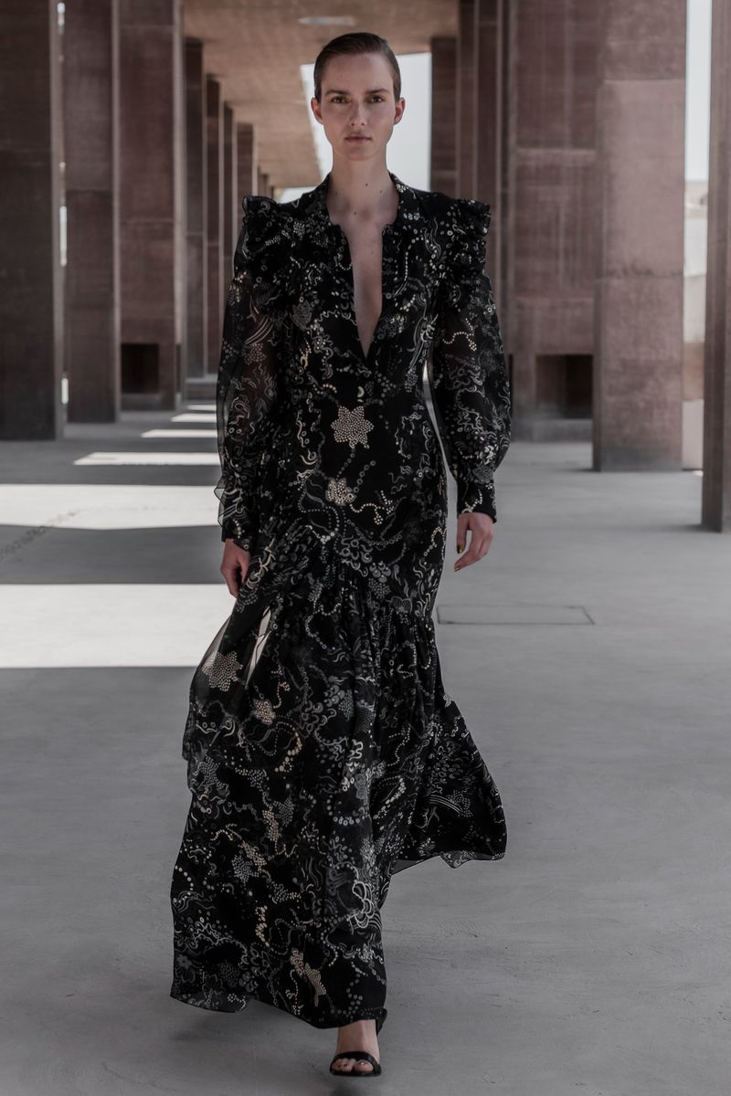 Fashion model walks along a concrete colonnade wearing a long black floral gown with puffy sleeves and sandals in an editorial fashion shot.