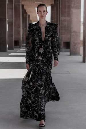 Fashion model walks along a concrete colonnade wearing a long black floral gown with puffy sleeves and sandals in an editorial fashion shot.