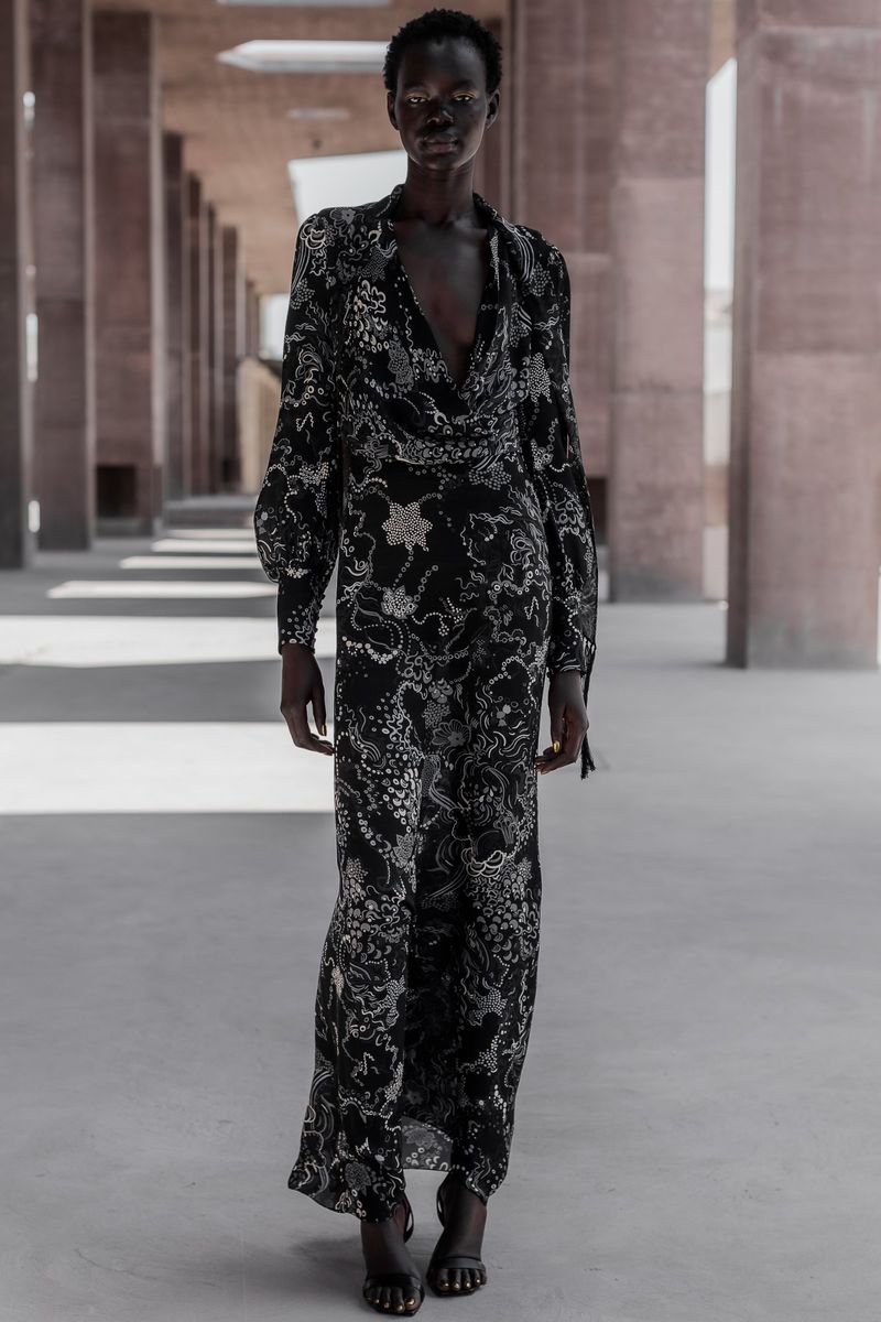 An adult female model with dark skin tone walks through an outdoor hallway wearing a black floral print gown during daytime.