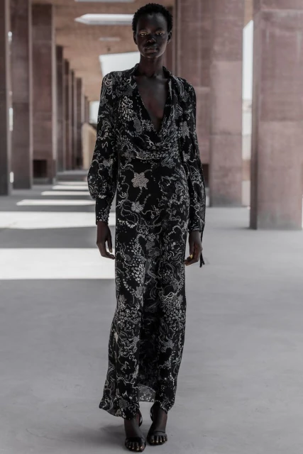 An adult female model with dark skin tone walks through an outdoor hallway wearing a black floral print gown during daytime.