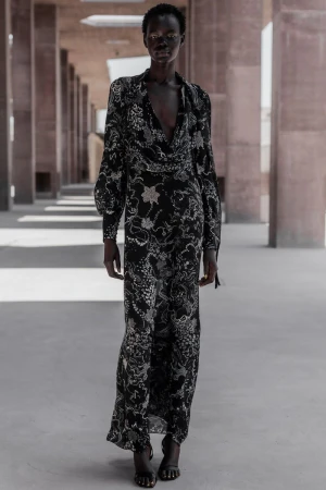 An adult female model with dark skin tone walks through an outdoor hallway wearing a black floral print gown during daytime.