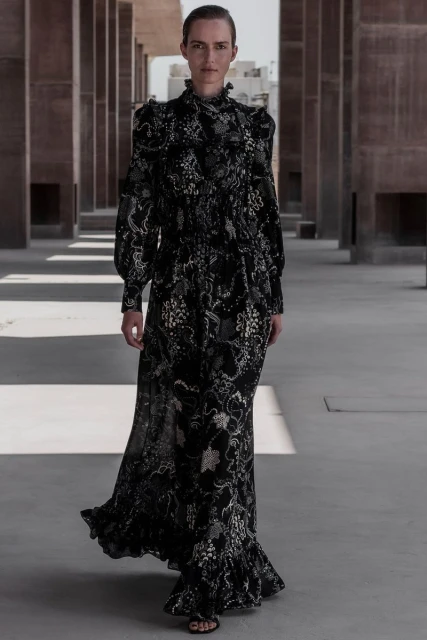 Elegant woman in a long-sleeve, floral maxi dress walking outdoors, with an eye-level view of a geometric, architectural background.