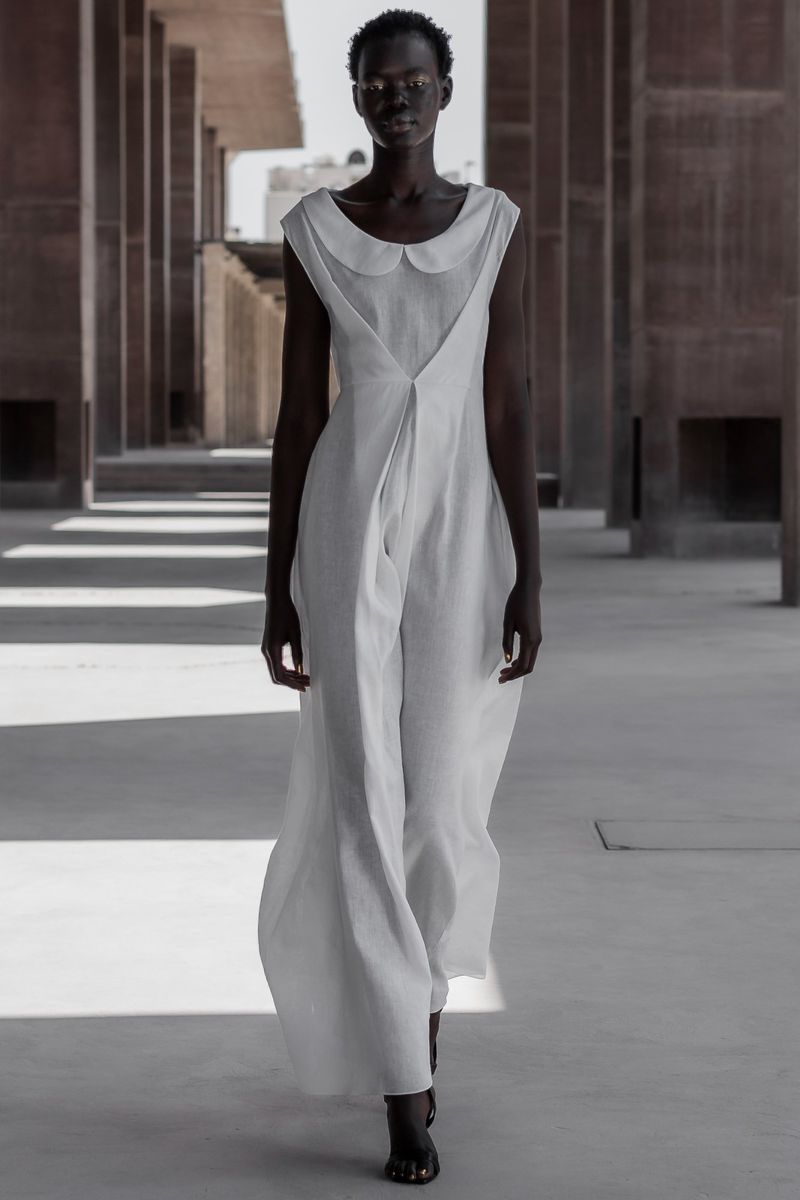 A slender, dark-skinned model in a flowing white jumpsuit confidently walks through a modern building with structured columns and diffused light.