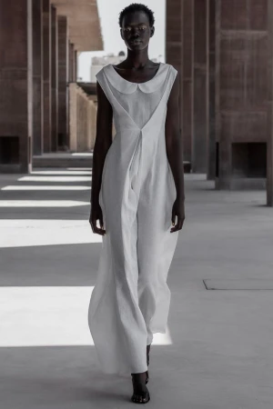A slender, dark-skinned model in a flowing white jumpsuit confidently walks through a modern building with structured columns and diffused light.