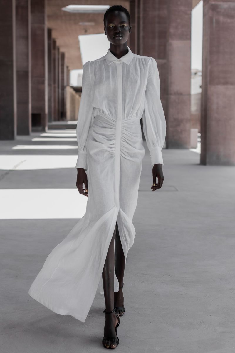 Elegant African woman in a white dress with a front slit, walking in a modern architectural space with strong lines and diffused natural light.