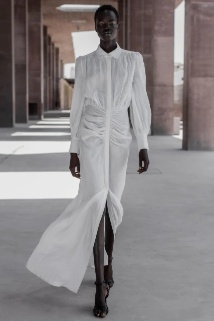 Elegant African woman in a white dress with a front slit, walking in a modern architectural space with strong lines and diffused natural light.